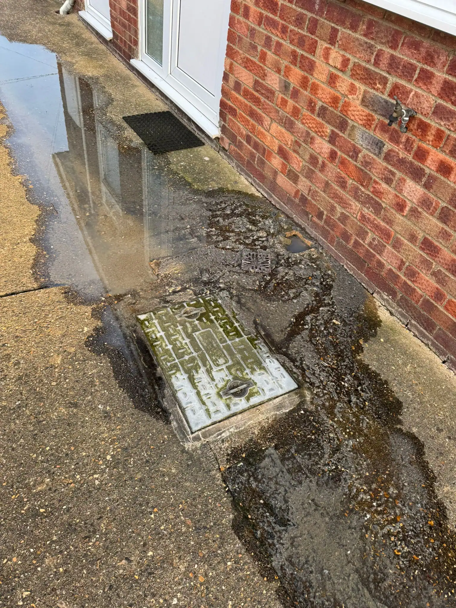 Emergency drain unblocking King's Lynn showing wastewater overflowing around a blocked drain inspection chamber outside a residential property.
